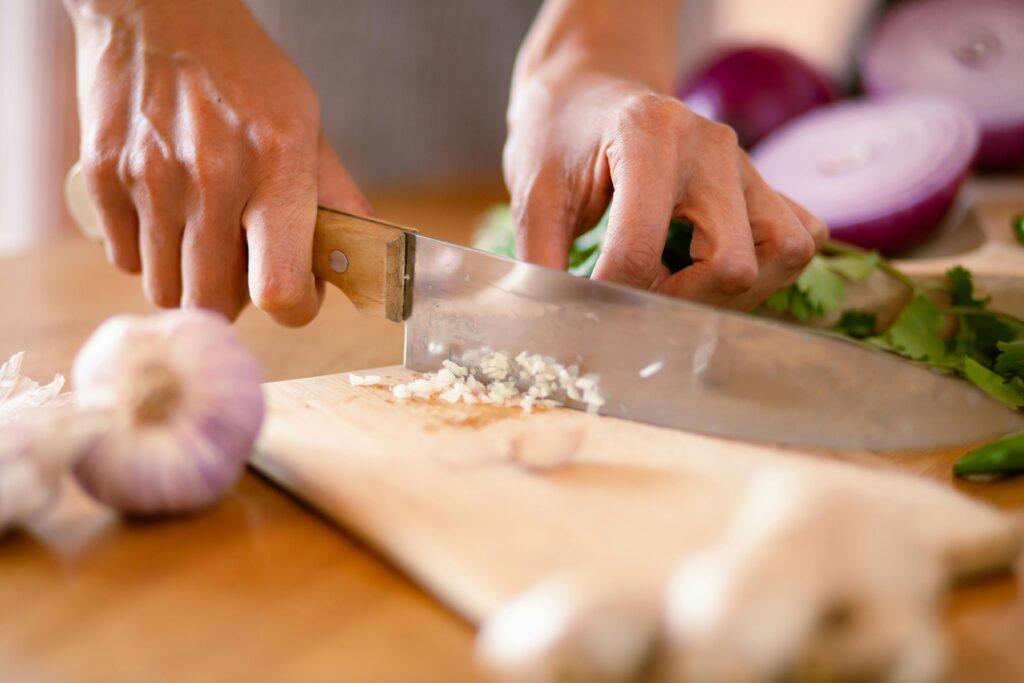 Hands chopping fresh garlic and onions on a wooden board in a kitchen setting.