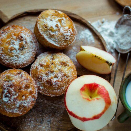 Top view of homemade apple muffins with sugar dusting, fresh apples, and a cup of milk on a wooden board.