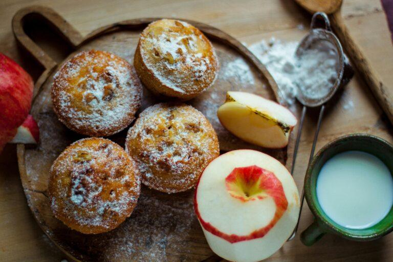 Top view of homemade apple muffins with sugar dusting, fresh apples, and a cup of milk on a wooden board.